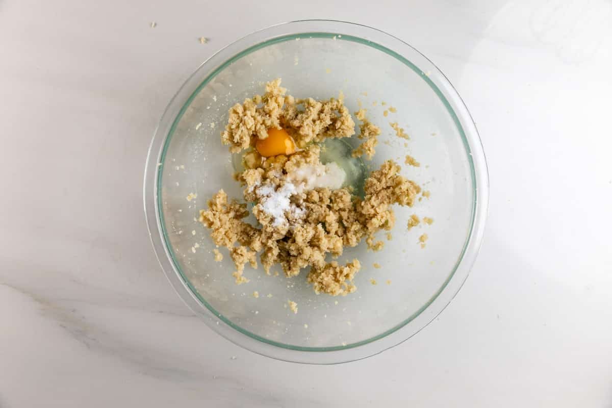 A glass mixing bowl with creamed butter and sugar, a cracked egg, and a pile of flour on a white countertop, ready to be mixed for baking.