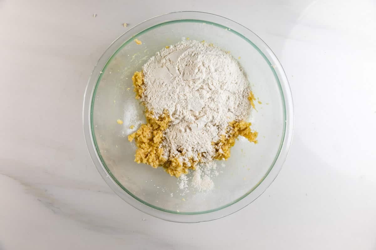 A glass mixing bowl containing creamed butter and sugar with a mound of flour on top, set on a white countertop, ready to be mixed for baking.