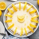 A lemon pie topped with whipped cream and lemon slices sits on a gray surface, surrounded by whole and sliced lemons. Nearby are two white plates with forks.