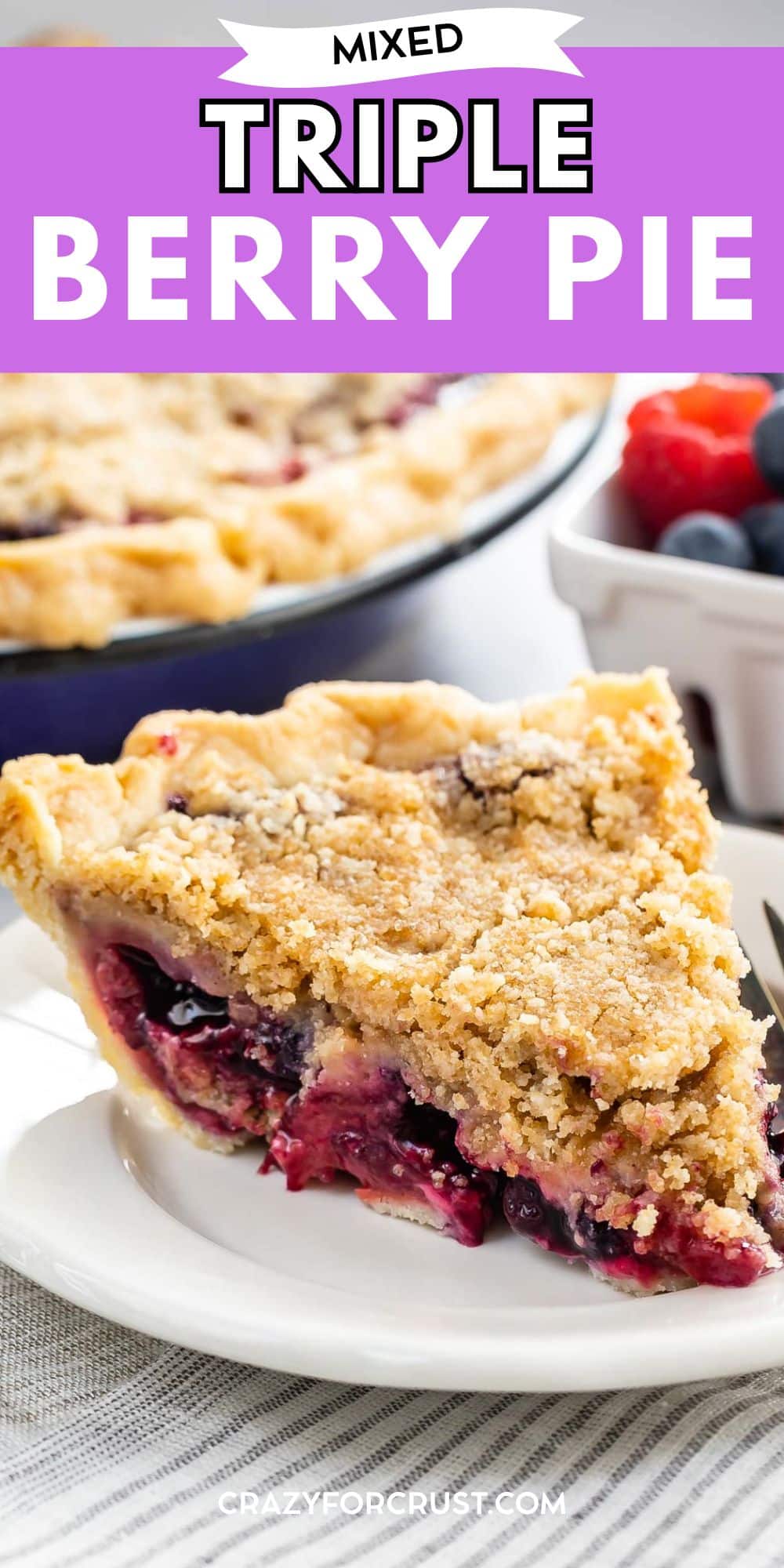 A slice of triple berry pie with a crumbly golden crust sits on a white plate, with fresh berries and the rest of the pie visible in the background. A fork rests beside the slice.