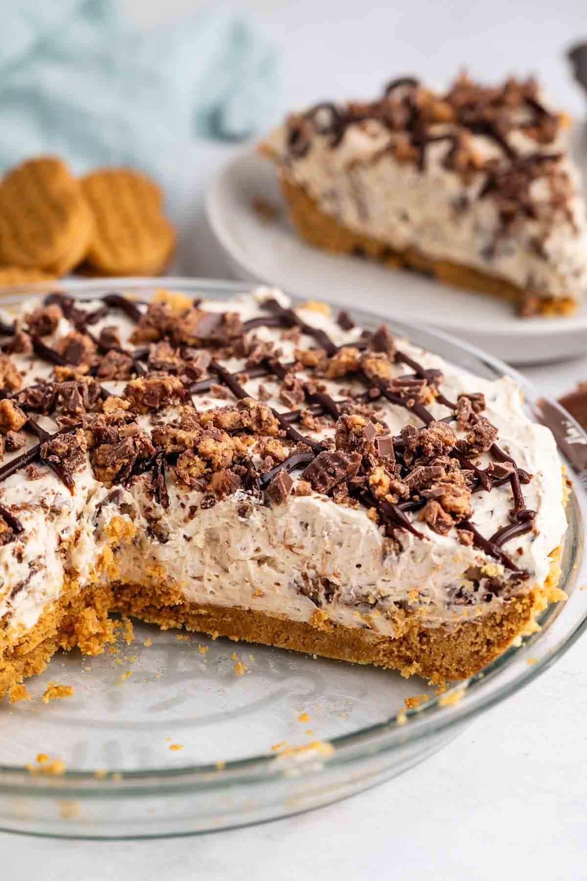 A creamy, no-bake pie with a graham cracker crust, topped with chocolate drizzle and crumbled cookies, sits in a glass pie dish. A slice has been removed and placed on a white plate in the background.