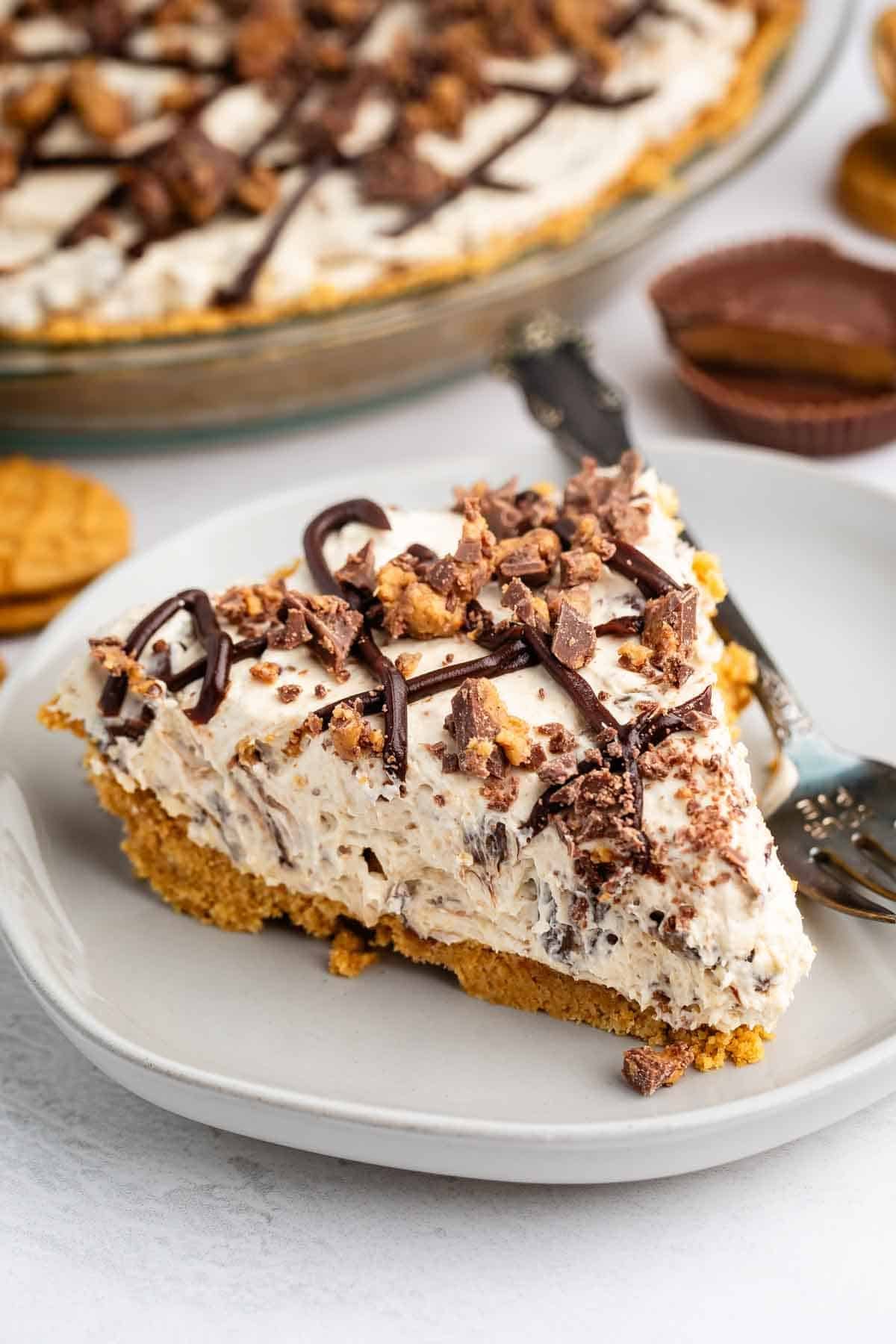 A slice of creamy pie with a crumbly crust sits on a plate, topped with chocolate drizzle and chopped peanut butter cups, with the rest of the pie and peanut butter cookies in the background.