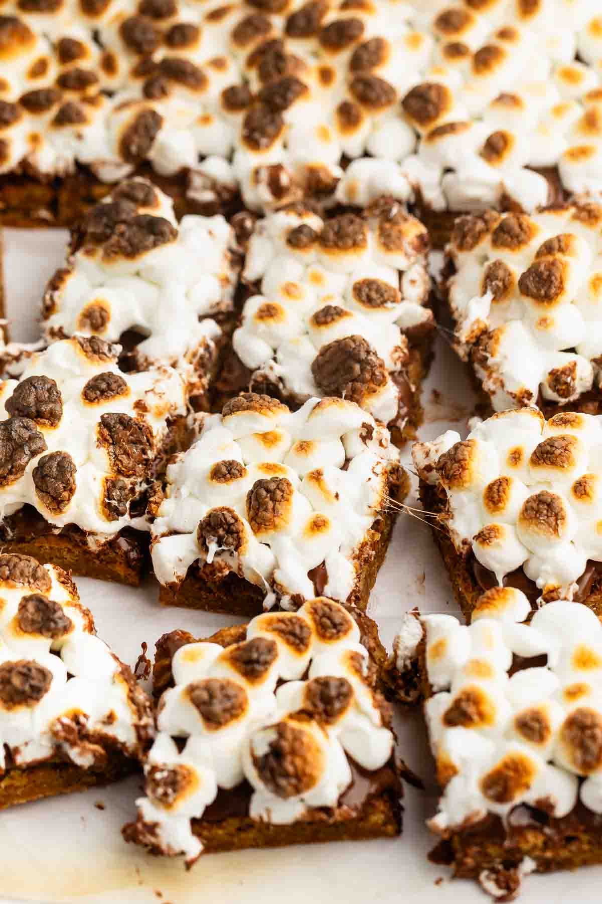 Smores bars cut into squares, topped with toasted marshmallows and chocolate chunks, arranged closely together on a parchment-lined surface.