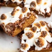 Close-up of dessert bars topped with toasted marshmallows and melted chocolate, with a golden brown base, arranged on a light surface. One bar is slightly lifted, showing the gooey texture.