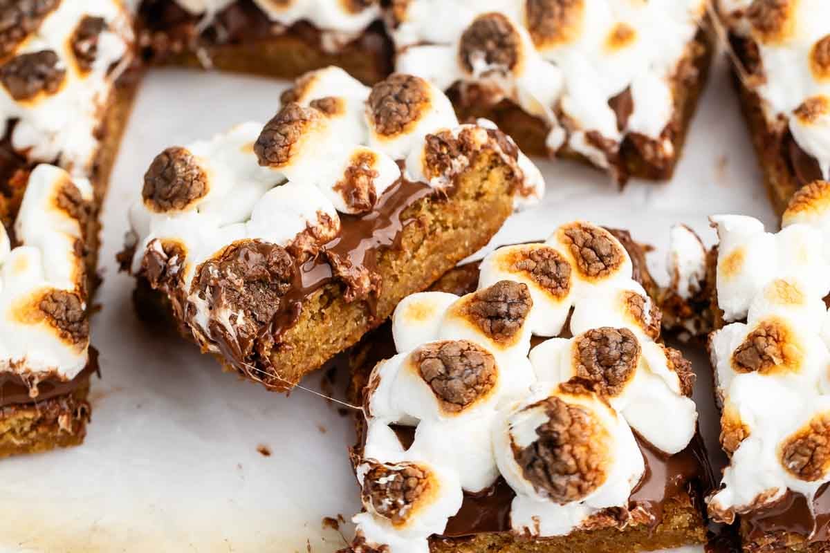 Close-up of dessert bars topped with toasted marshmallows and melted chocolate, with a golden brown base, arranged on a light surface. One bar is slightly lifted, showing the gooey texture.