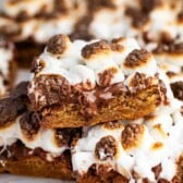 Close-up of gooey, golden-brown smores bars topped with toasted marshmallows, melted chocolate, and pieces of graham crackers, stacked on top of each other.
