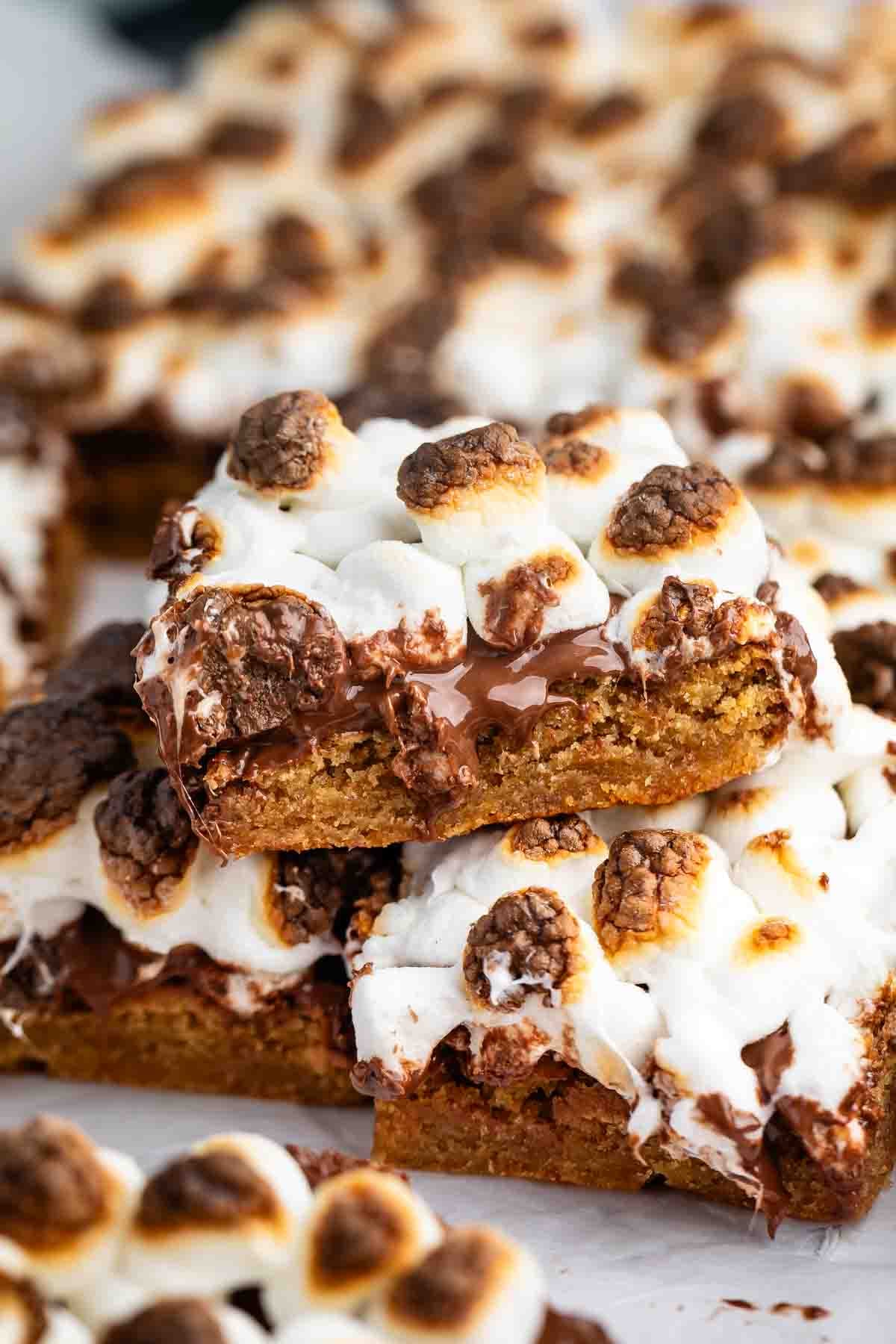 Close-up of gooey, golden-brown smores bars topped with toasted marshmallows, melted chocolate, and pieces of graham crackers, stacked on top of each other.