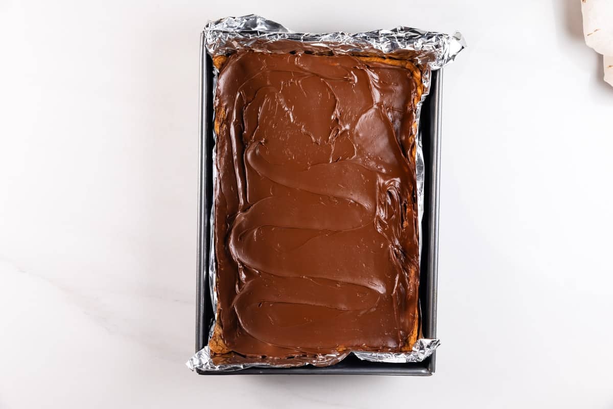 A rectangular baking pan lined with foil holds a dessert topped with a smooth, glossy layer of chocolate. The pan is placed on a white surface.