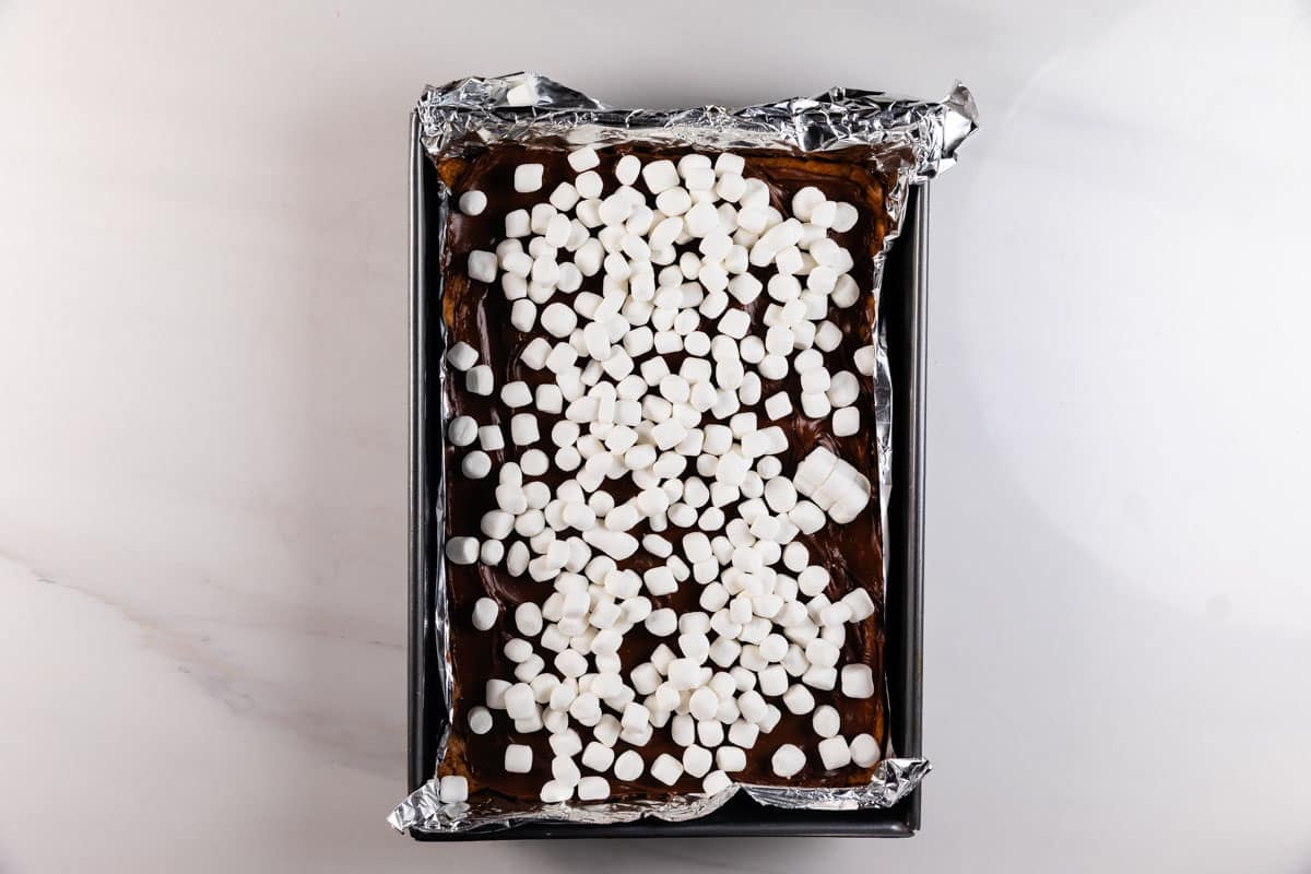 A rectangular baking pan lined with foil contains a chocolate dessert topped with a layer of mini marshmallows, viewed from above on a light countertop.