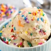 Three scoops of vanilla ice cream topped with colorful rainbow sprinkles in a light blue bowl, with a blurred dish of sprinkles and two spoons in the background.