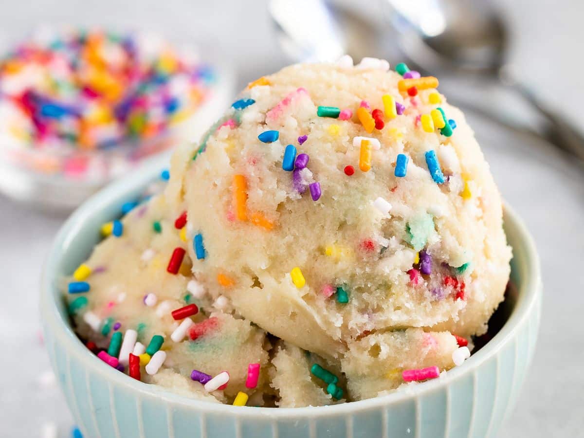 Three scoops of vanilla ice cream topped with colorful rainbow sprinkles in a light blue bowl, with a blurred dish of sprinkles and two spoons in the background.