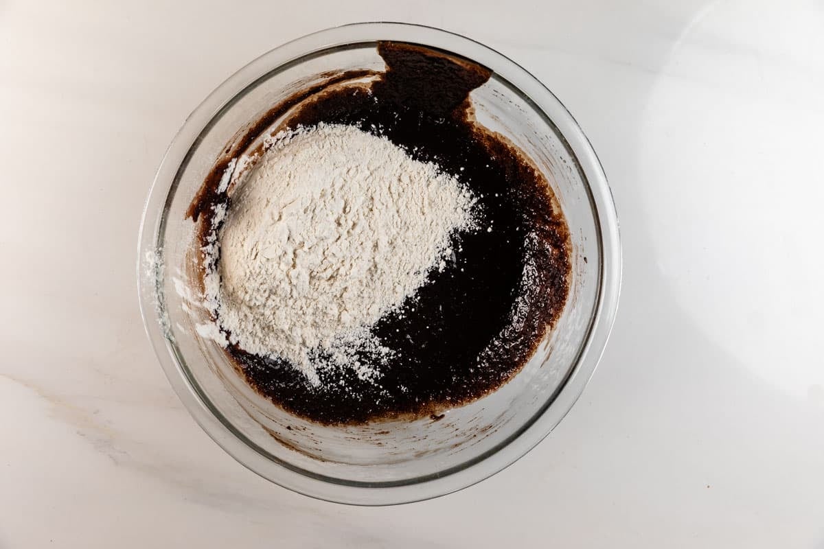 A glass bowl containing brownie batter with a pile of white flour on top, placed on a white countertop.