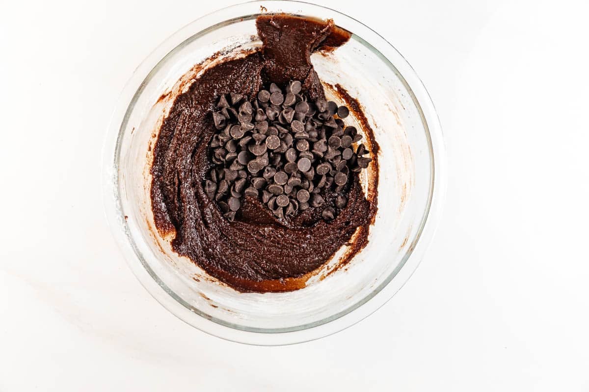 A glass bowl filled with chocolate brownie batter and topped with a pile of chocolate chips, viewed from above on a white surface.
