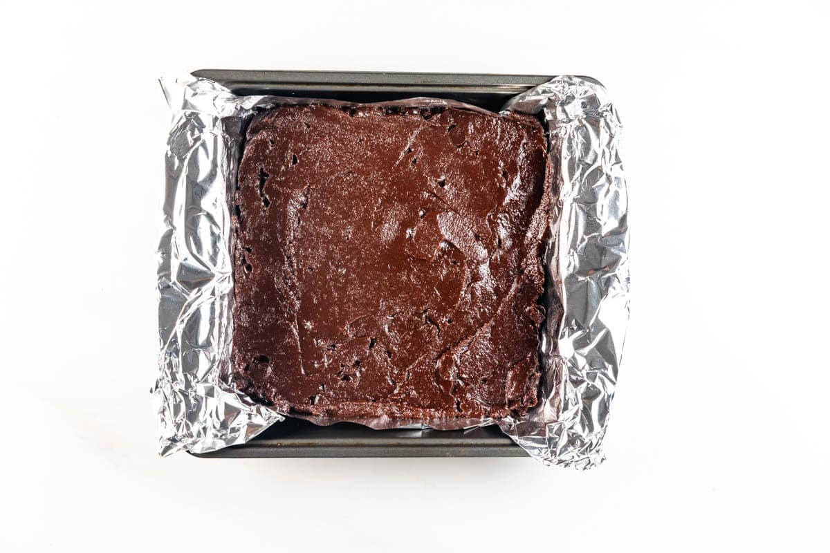 A square baking pan lined with crinkled aluminum foil, filled with unbaked chocolate brownie batter, is shown from above on a white background.
