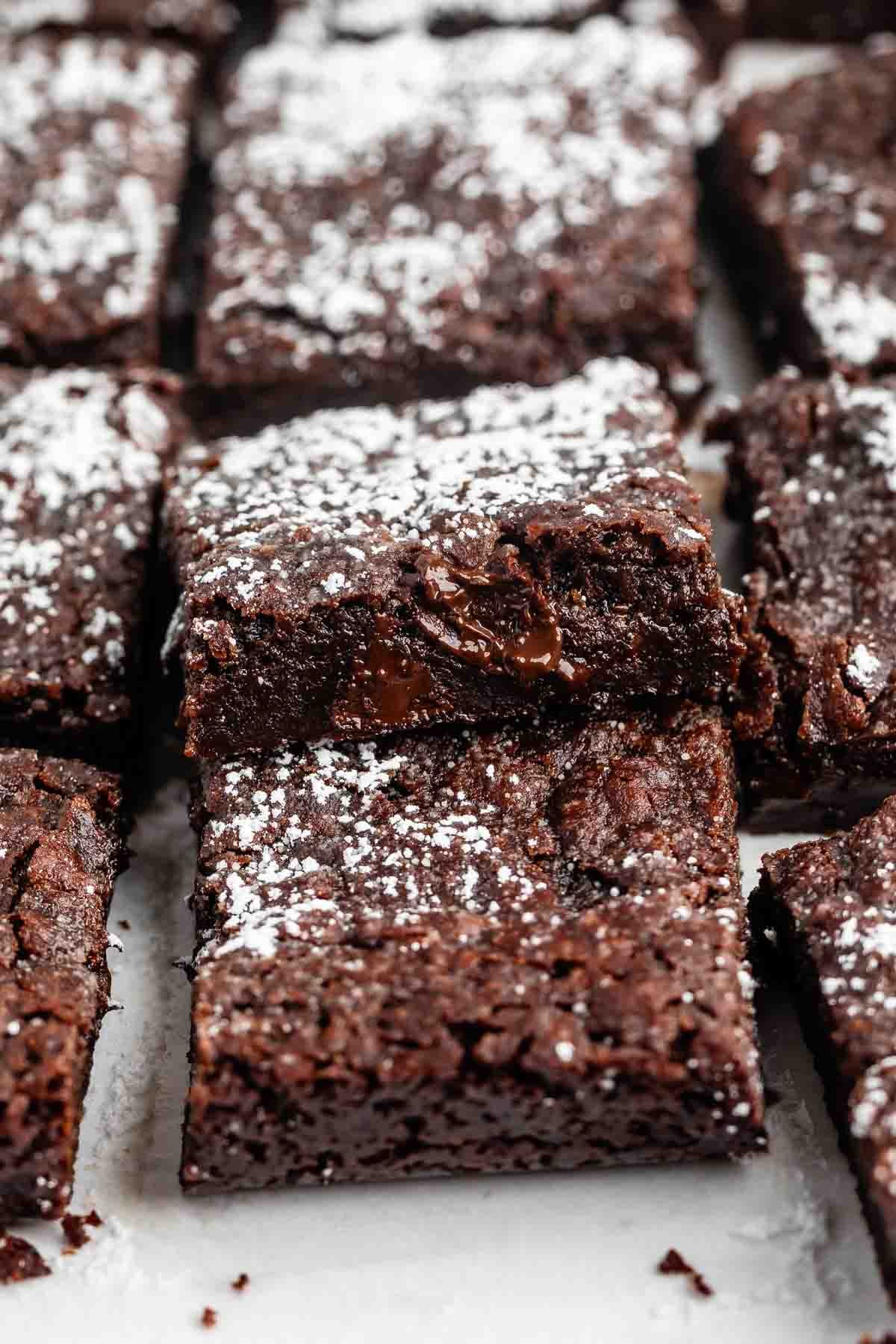 A close-up of several chocolate brownies topped with powdered sugar, with one brownie slightly tilted to reveal a gooey, melted chocolate center.