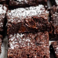 Close-up of rich, fudgy chocolate brownies topped with a light dusting of powdered sugar. The brownies are cut into squares and stacked, showing a moist and gooey texture with chunks of chocolate inside.