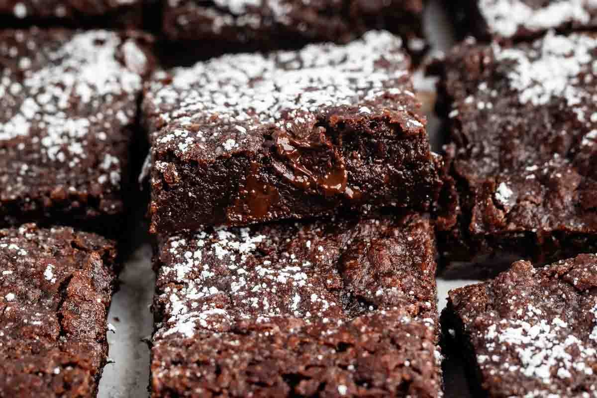 Close-up of rich, fudgy chocolate brownies topped with a light dusting of powdered sugar. The brownies are cut into squares and stacked, showing a moist and gooey texture with chunks of chocolate inside.