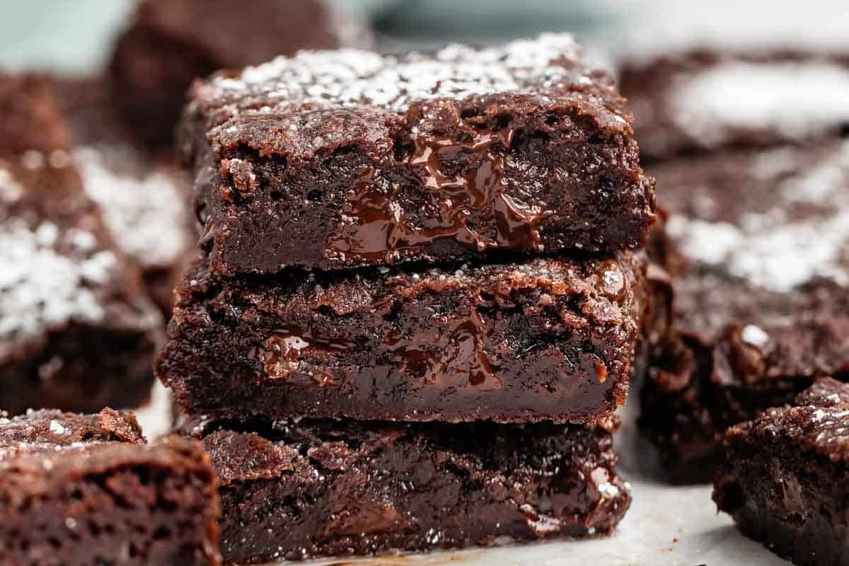 A close-up of three rich, fudgy chocolate brownies stacked on top of each other, with melted chocolate inside and a light dusting of powdered sugar on top.