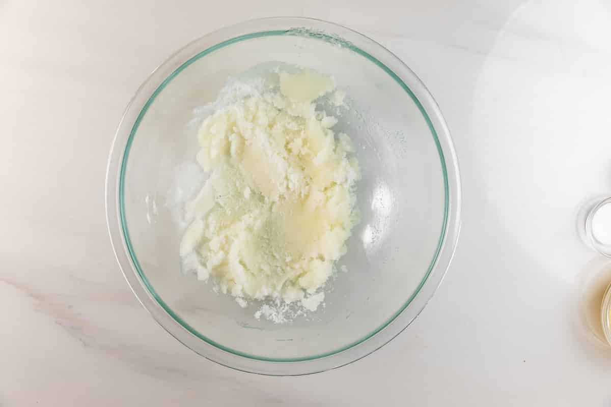 A glass mixing bowl on a white countertop containing creamed butter and sugar, partially mixed together. A small bowl with liquid is partially visible at the bottom right corner.