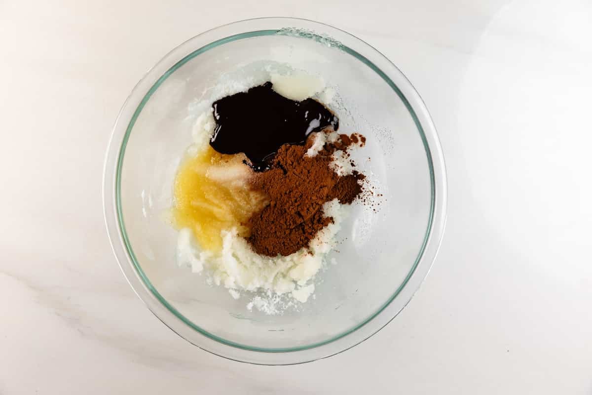 A glass mixing bowl containing white granulated substances, applesauce, dark syrup or molasses, and cocoa powder on a white countertop.
