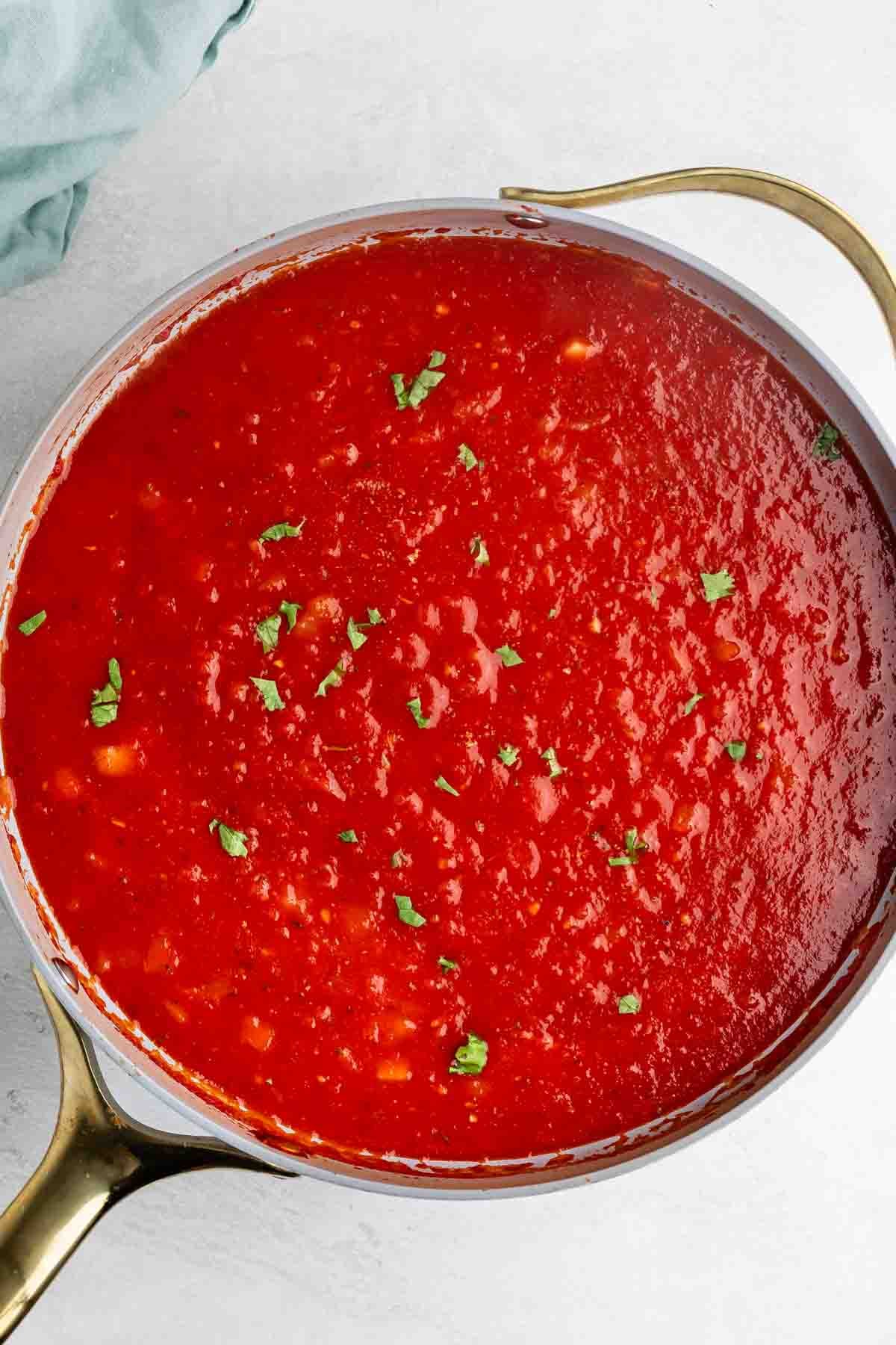 A saucepan filled with bright red tomato sauce, garnished with small pieces of fresh green herbs, sits on a light surface with a gold handle visible.