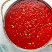 A pot filled with red marinara sauce, garnished with chopped fresh herbs, sits on a white surface next to a light blue cloth.