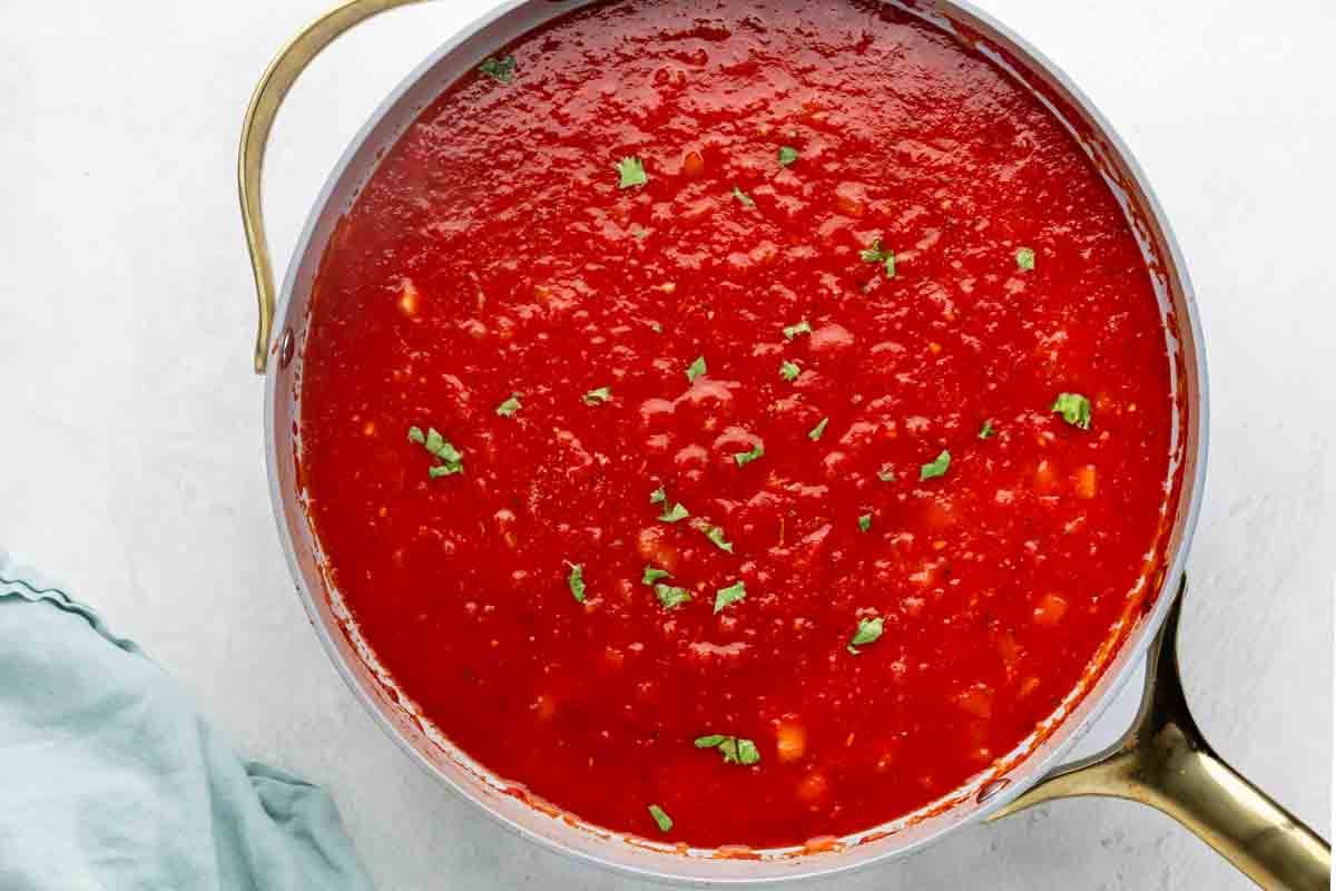 A pot filled with red marinara sauce, garnished with chopped fresh herbs, sits on a white surface next to a light blue cloth.