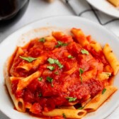 A white plate filled with penne pasta topped with marinara sauce and garnished with chopped fresh basil. A block of cheese and a glass of red wine are visible in the background.