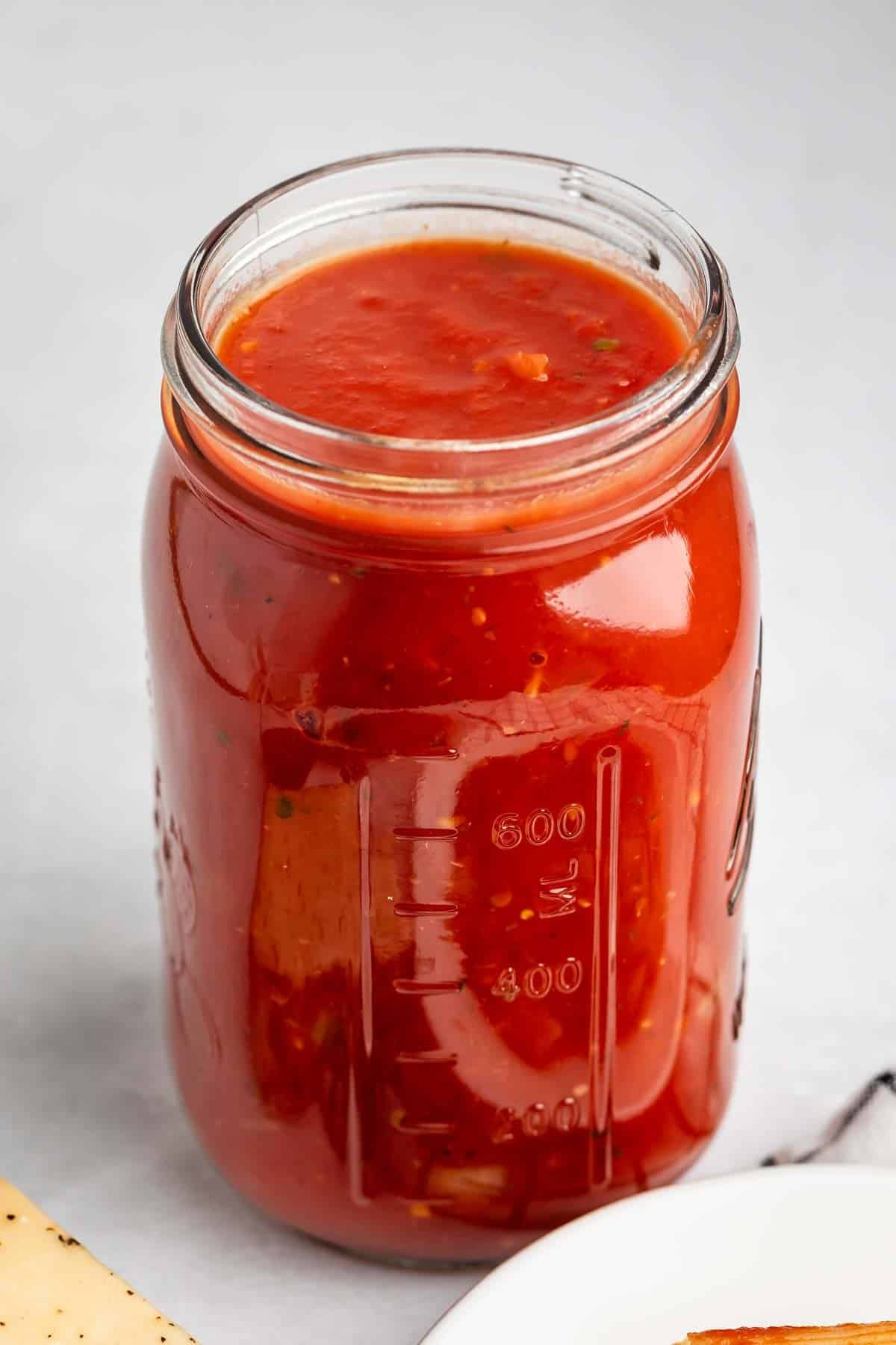 A glass mason jar filled with red marinara sauce sits on a light gray surface. The jar is nearly full, and the sauce has visible herbs and small pieces of tomato.