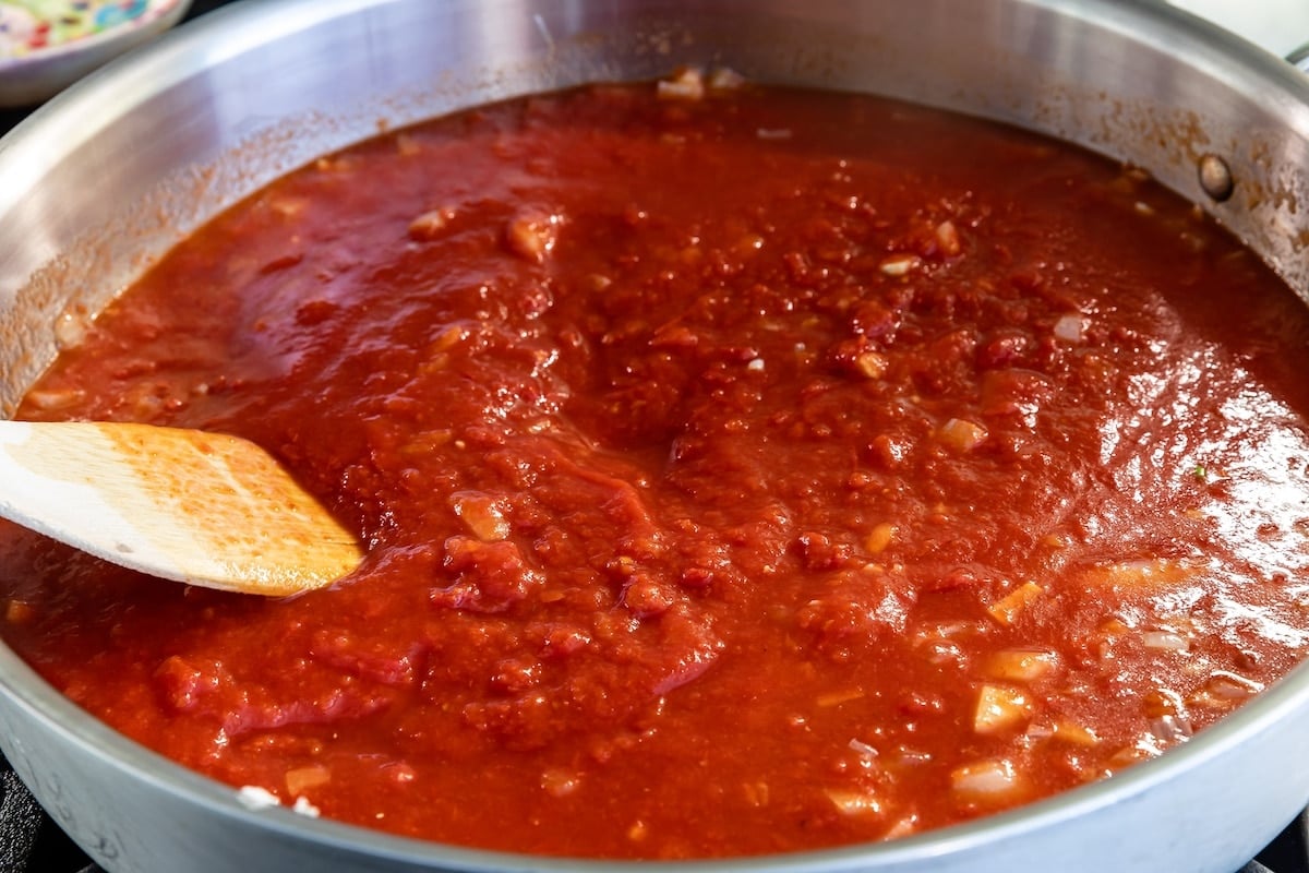 A close-up of a pot filled with simmering tomato sauce, with visible chunks of tomatoes and onions. A wooden spoon rests in the sauce, partially coated with the red mixture.