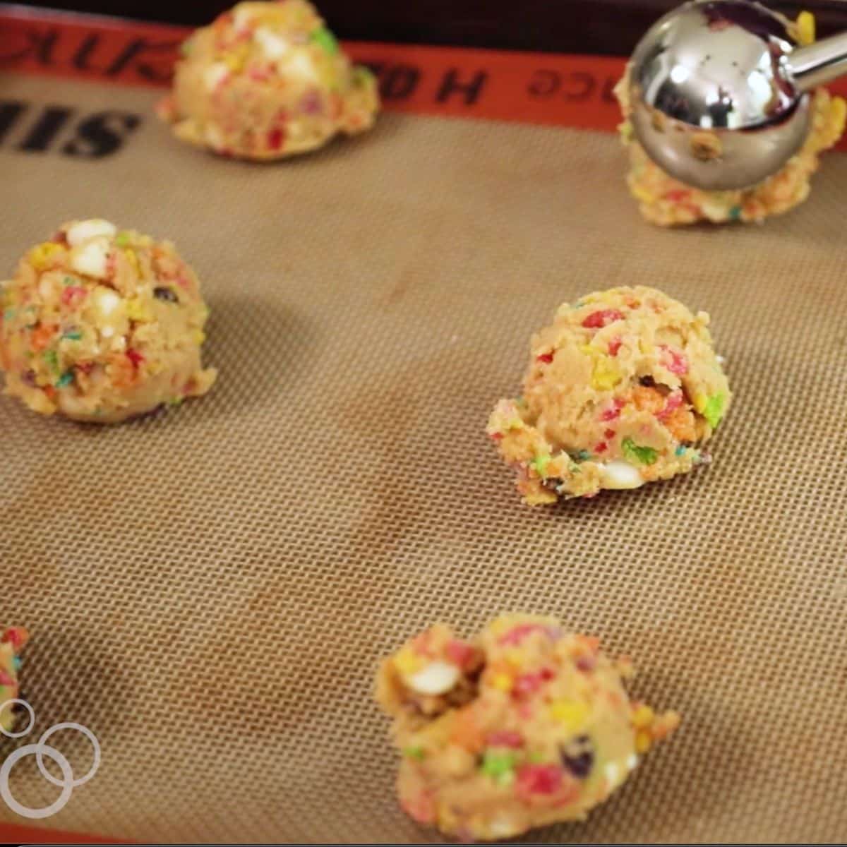 Scoops of colorful cookie dough with cereal pieces on a silicone baking mat, ready for baking. A metal cookie scoop is visible in the top right corner.