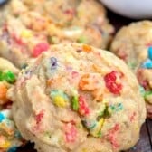 A close-up of a soft, thick cookie filled with colorful cereal pieces, resting on a dark wooden surface with more cookies and cereal pieces nearby.