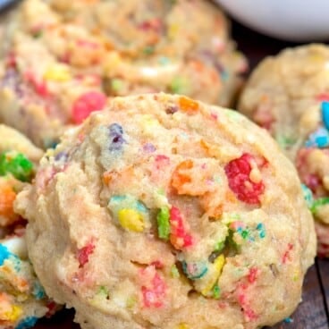 A close-up of a soft, thick cookie filled with colorful cereal pieces, resting on a dark wooden surface with more cookies and cereal pieces nearby.