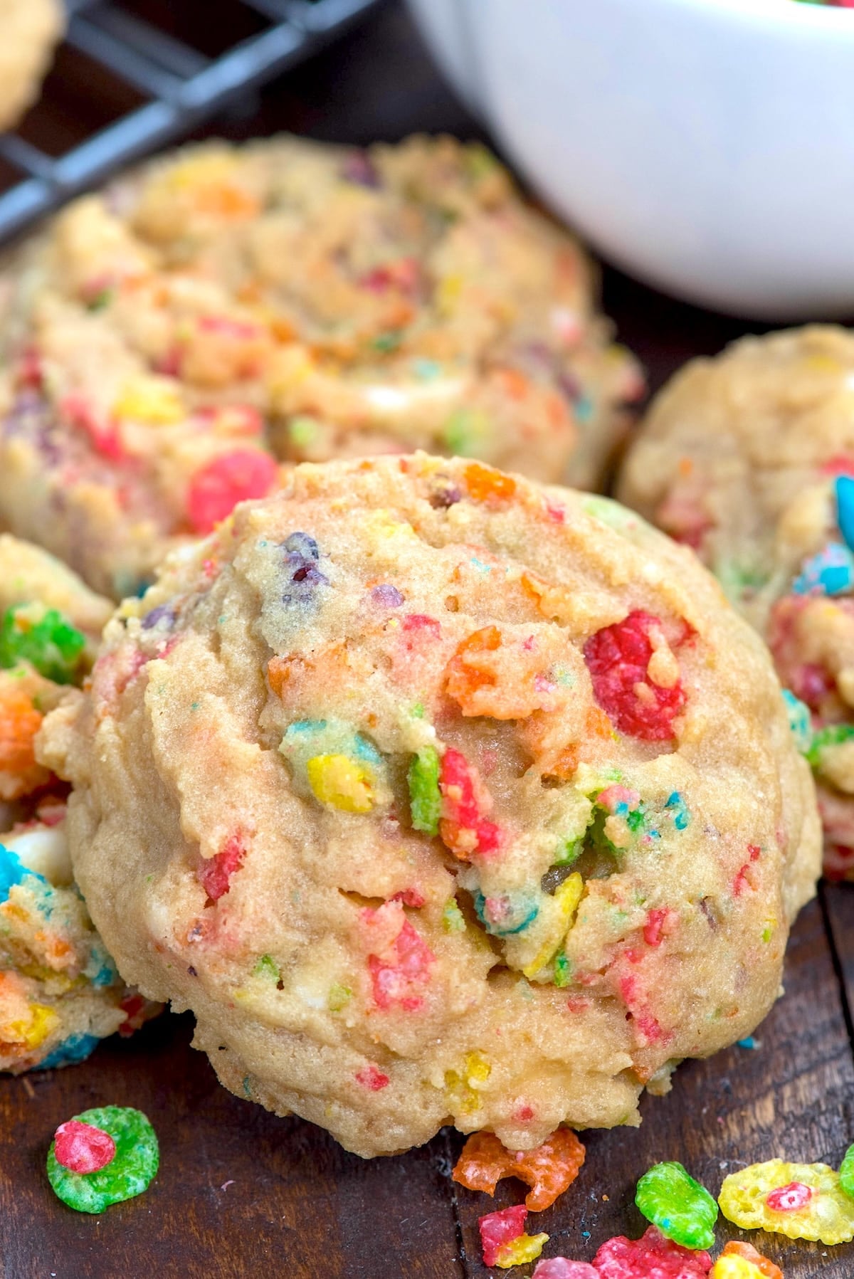 A close-up of a soft, thick cookie filled with colorful cereal pieces, resting on a dark wooden surface with more cookies and cereal pieces nearby.