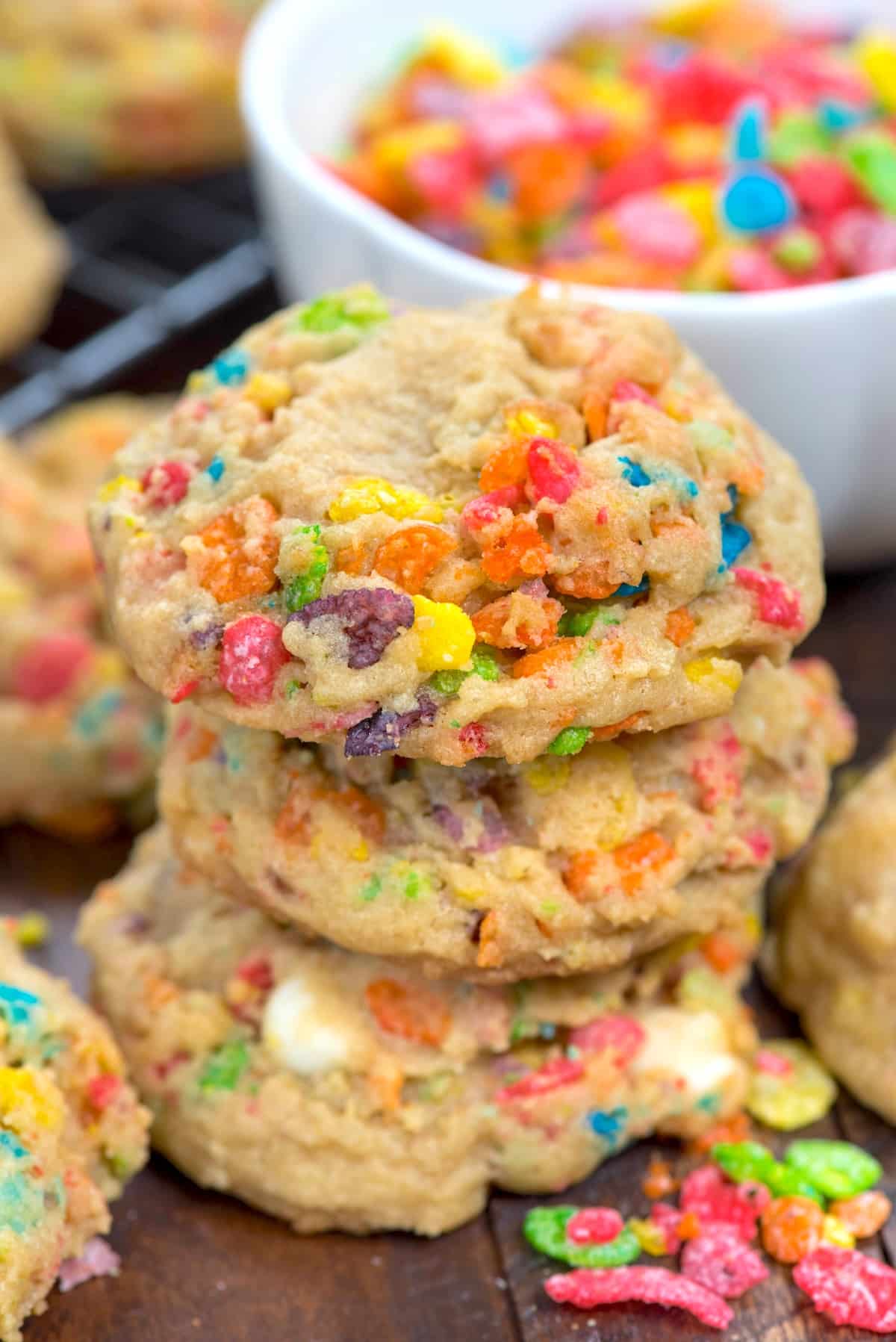 A close-up of three cookies stacked on top of each other, filled with colorful Fruity Pebbles cereal pieces, with more cookies and a bowl of Fruity Pebbles in the background.