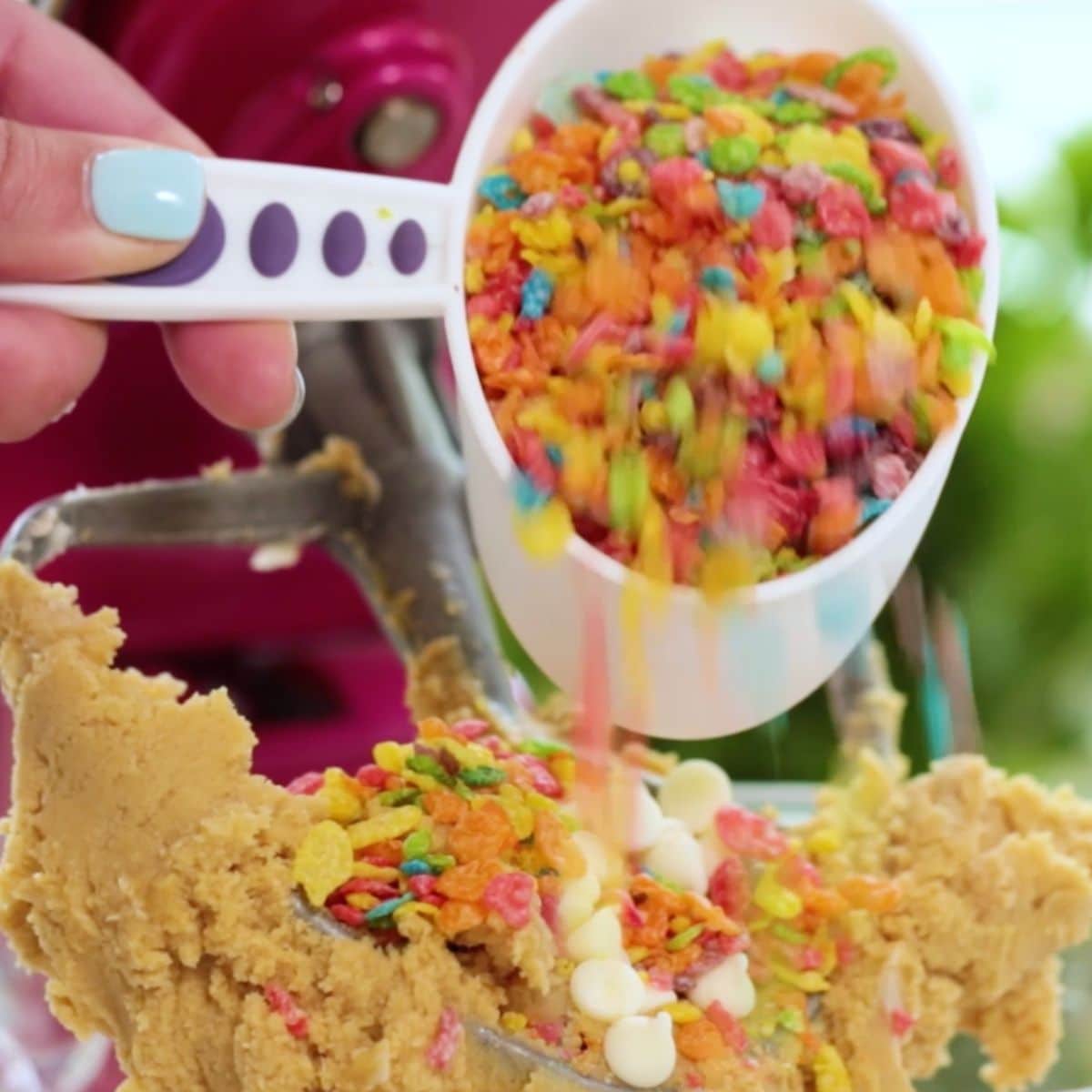 A hand pours colorful Fruity Pebbles cereal from a measuring cup into cookie dough with white chocolate chips, mixing with a stand mixer in the background.