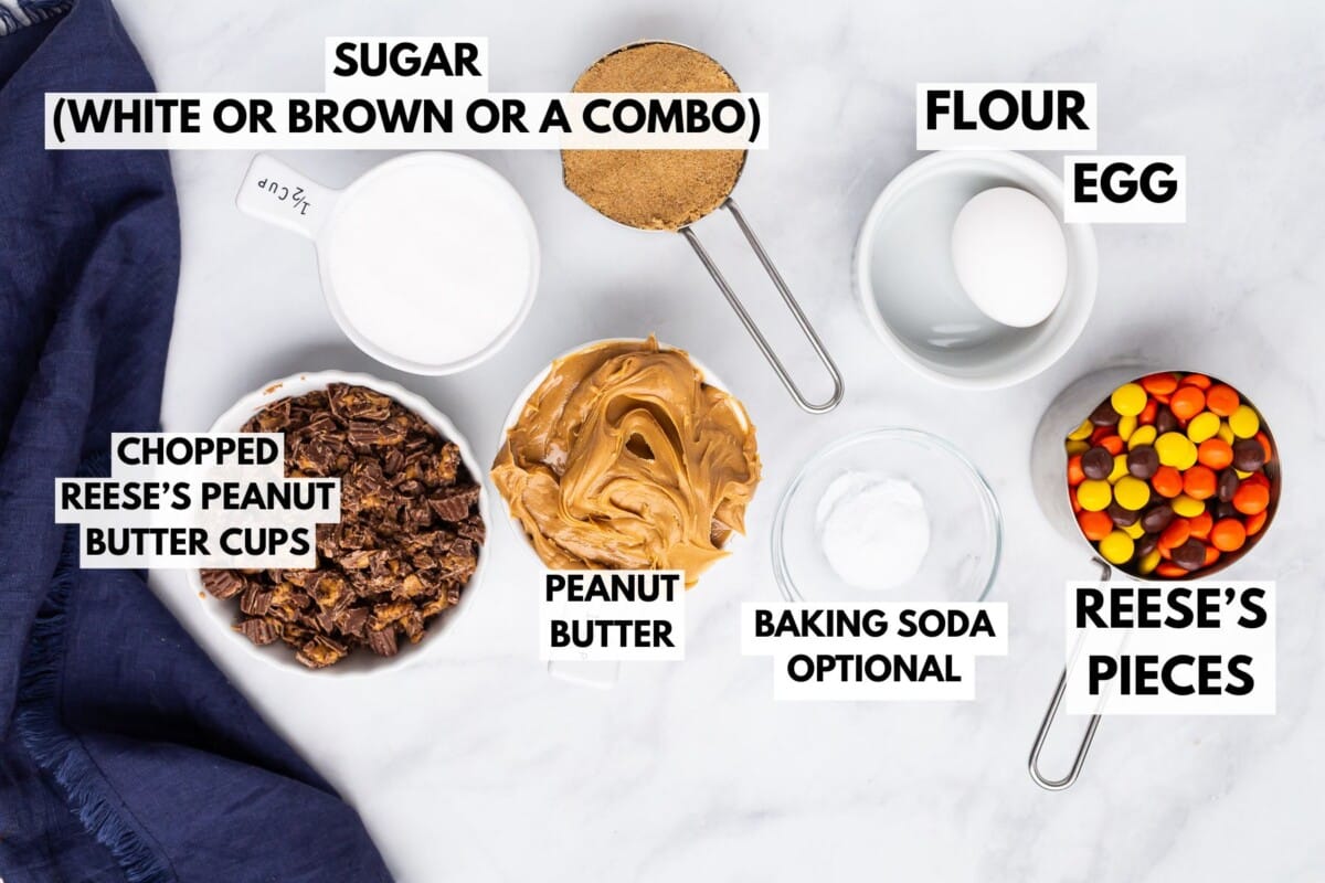 Ingrédients de cuisson sur une surface blanche : un bol de sucre, une tasse à mesurer de farine, un œuf, des coupes de beurre de cacahuète Reese, des coupes de beurre de cacahuète Reese, du bicarbonate de soude et des morceaux de Reese, étiquetés avec du texte.