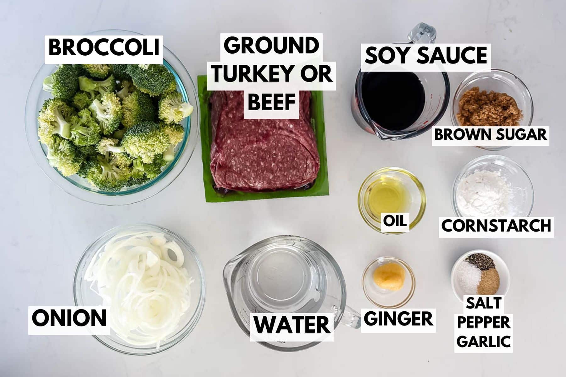 Ingredients for a stir fry arranged on a white surface: bowls of broccoli, sliced onion, ground turkey or beef, soy sauce, brown sugar, oil, cornstarch, ginger, water, salt, pepper, and garlic, all labeled.