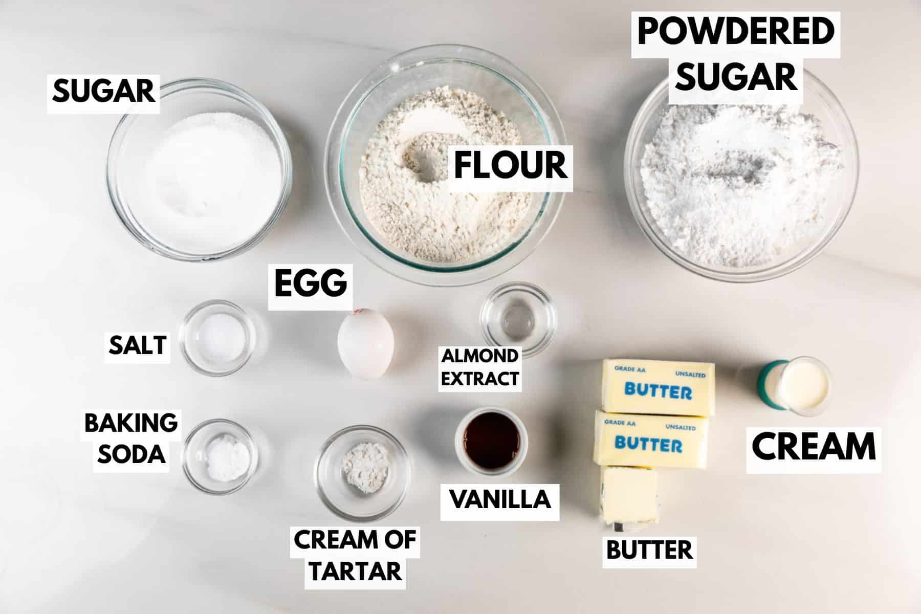 Meilleures barres de biscuits au sucre – Fou de croûte 6 Vue de haut en bas de divers ingrédients de cuisson étiquetés dans des bols et des récipients, notamment le sucre, le sucre en poudre, la farine, le sel, le bicarbonate de soude, la crème de tartre, l'extrait d'amande, la vanille, les œufs, la crème et le beurre sur une surface blanche.