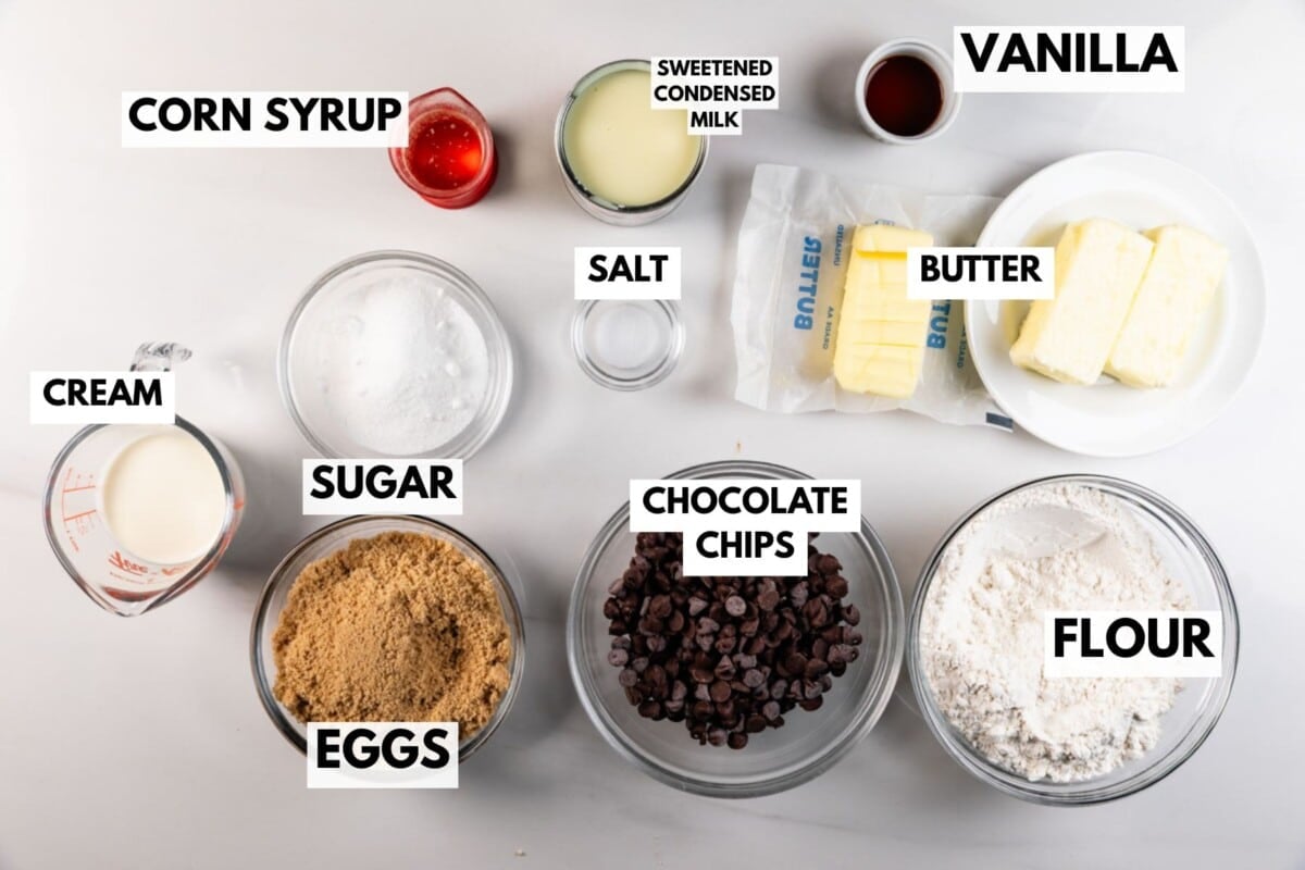 Overhead view of various baking ingredients in bowls and plates, each labeled: corn syrup, sweetened condensed milk, vanilla, cream, sugar, salt, butter, eggs, chocolate chips, and flour.