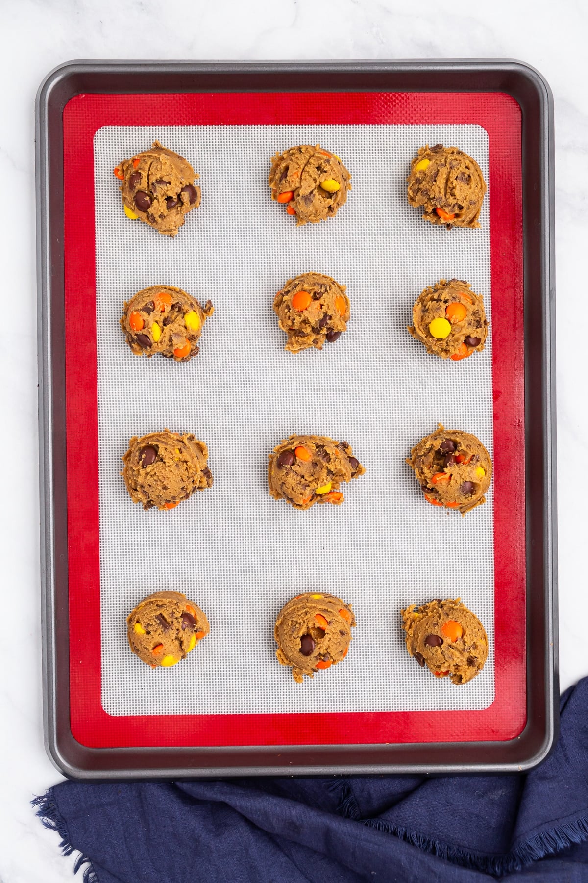 Douze boules de pâte à biscuits enrobée de chocolat coloré, uniformément espacées sur un tapis de cuisson en silicone sur une plaque à pâtisserie, prêtes à cuire. Un tissu bleu foncé est partiellement visible dans le coin inférieur.