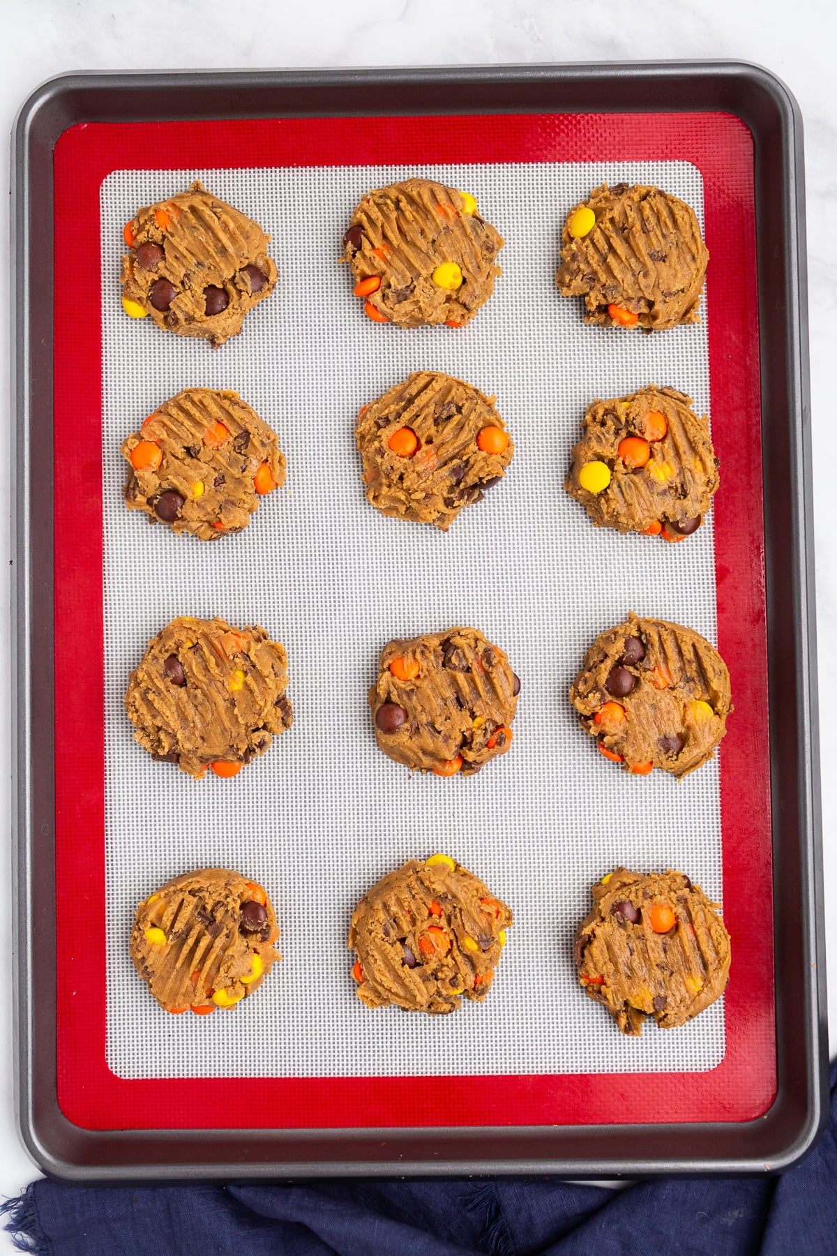 Une plaque à pâtisserie contenant douze biscuits non cuits contenant du chocolat et des bonbons à l'orange, disposés en rangées sur un tapis en silicone blanc, prêts à cuire.