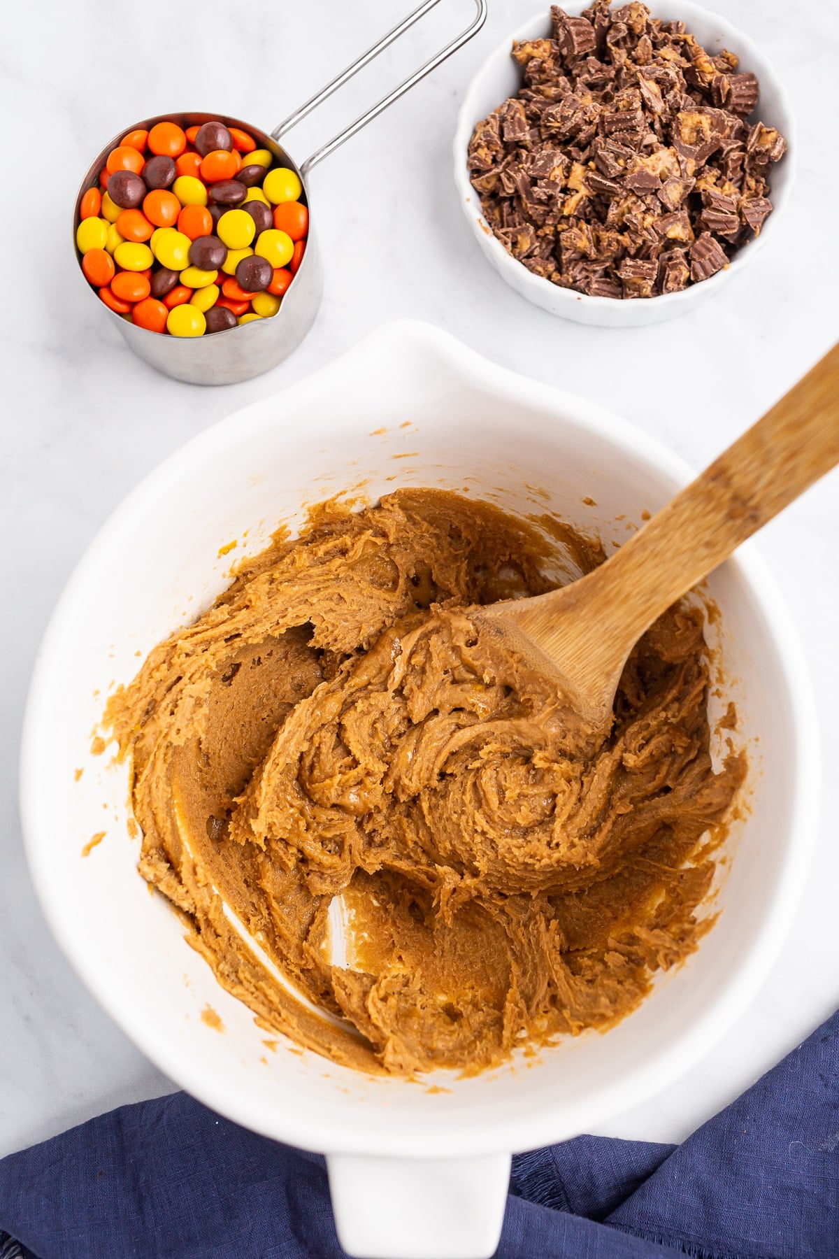 Un bol à mélanger blanc rempli de pâte à biscuits et une cuillère en bois, à côté d'un bol de bonbons au chocolat hachés et d'une tasse à mesurer avec des morceaux colorés de Reese sur une surface claire.