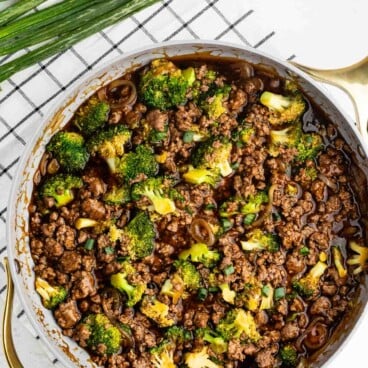 A skillet filled with ground beef, broccoli florets, and sliced green onions in a dark sauce, placed on a checkered cloth with fresh green onions beside it.