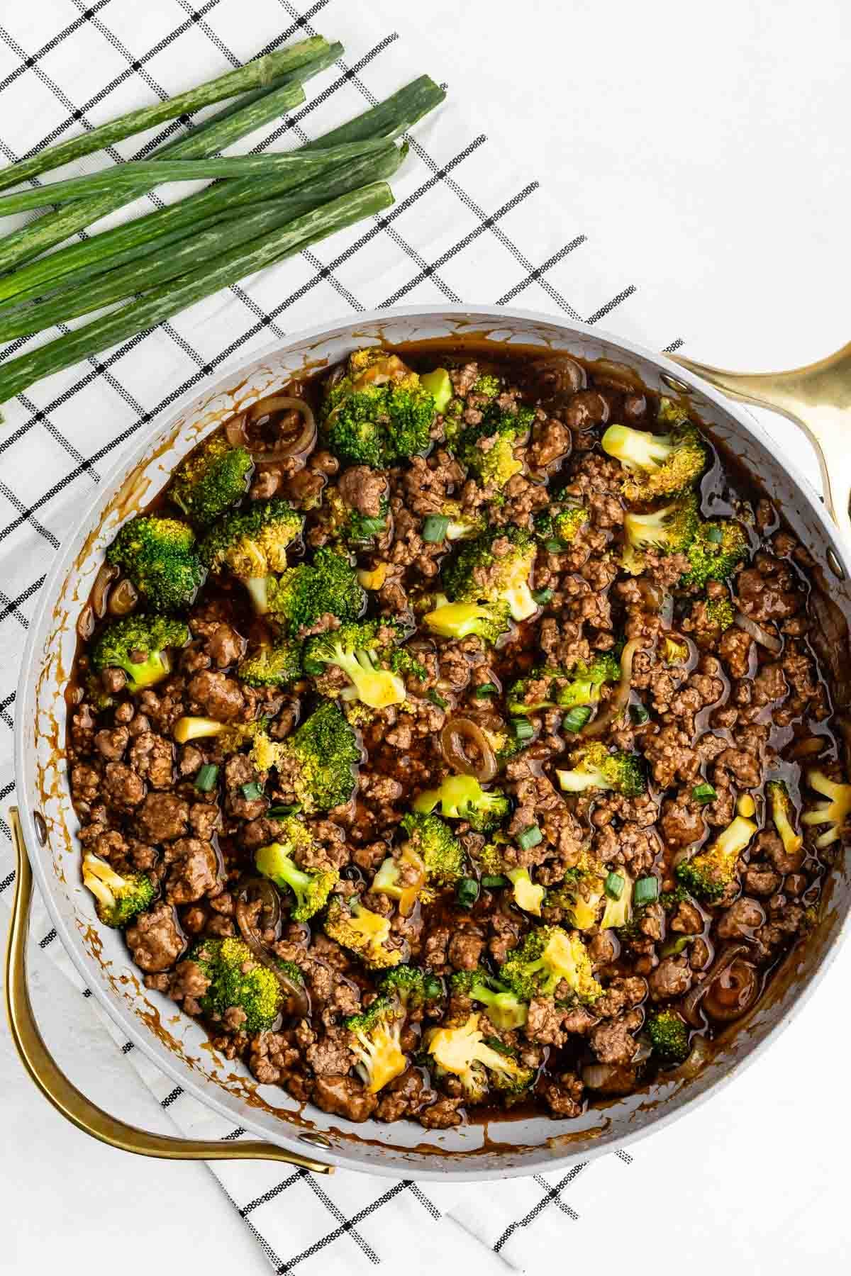 A skillet filled with ground beef, broccoli florets, and sliced green onions in a dark sauce, placed on a checkered cloth with fresh green onions beside it.