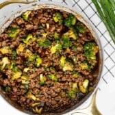A large skillet filled with cooked ground beef, broccoli florets, and a dark savory sauce, garnished with sliced green onions, sits on a checkered cloth next to a bunch of fresh green onions.