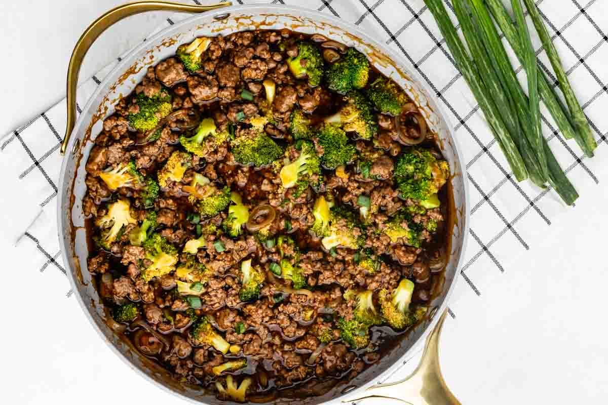 A large skillet filled with cooked ground beef, broccoli florets, and a dark savory sauce, garnished with sliced green onions, sits on a checkered cloth next to a bunch of fresh green onions.