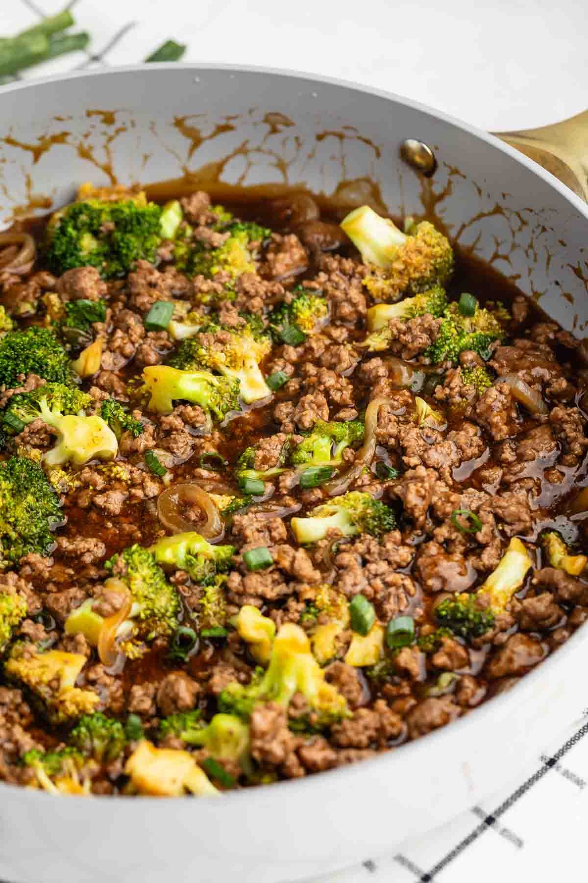 A skillet filled with cooked ground beef, broccoli florets, sliced onions, and green onions in a brown savory sauce.