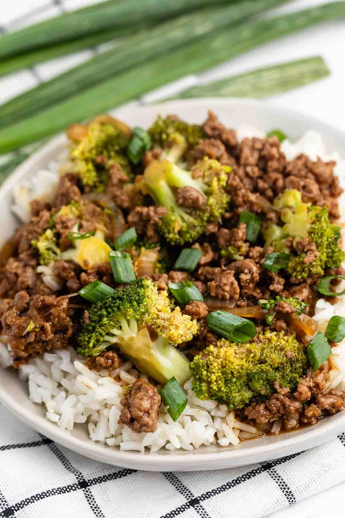 A plate of white rice topped with cooked ground beef, broccoli florets, and chopped green onions, with fresh green onions in the background.