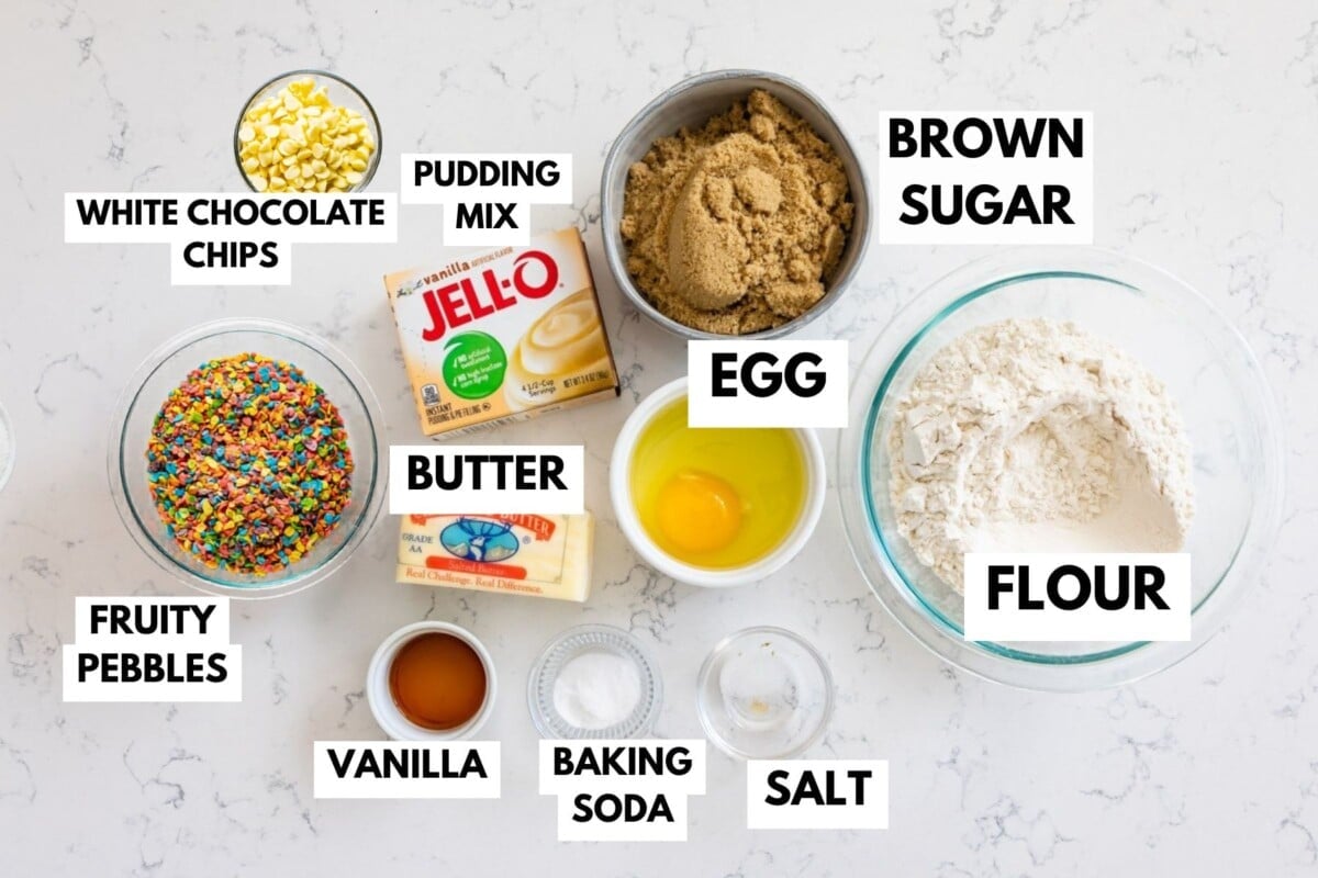 Top-down view of baking ingredients labeled: white chocolate chips, pudding mix, brown sugar, egg, flour, fruity pebbles cereal, butter, vanilla, baking soda, and salt on a white surface.